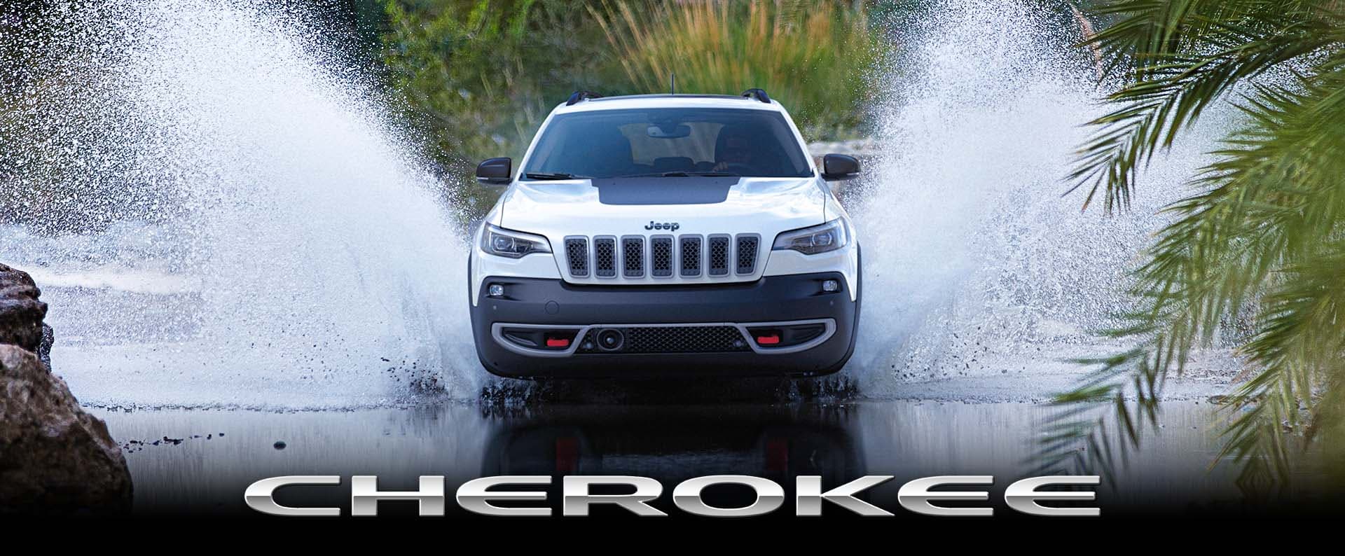 A head-on angle of a white 2023 Jeep Cherokee Trailhawk fording a stream off-road, with water splashing well above the top of the vehicle. Cherokee.