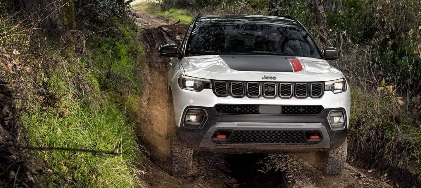 A head-on angle of a 2024 Jeep Compass Trailhawk being driven off-road through a narrow trail with thick growth on both sides.