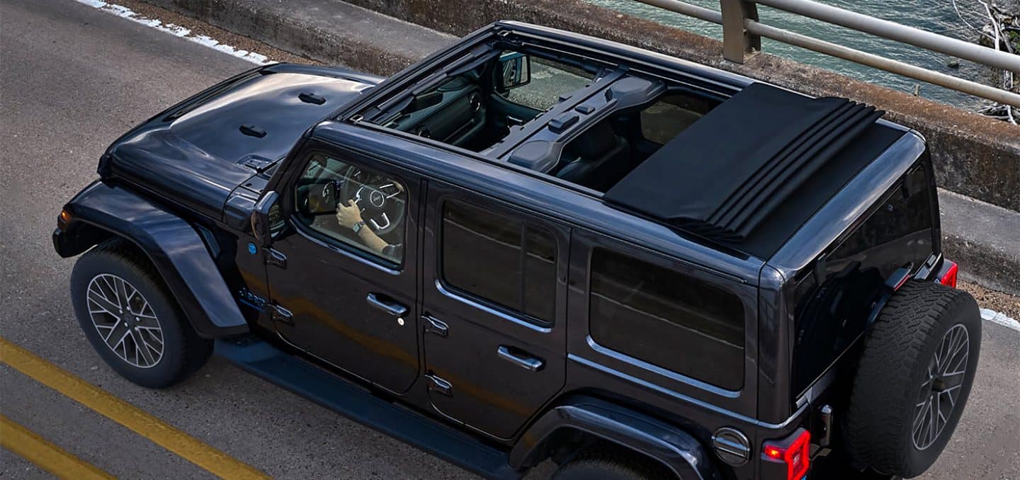 Display An angle from above of a black 2024 Jeep Wrangler High Altitude 4xe with its Sky One-Touch power sunroof fully open, being driven on a highway.