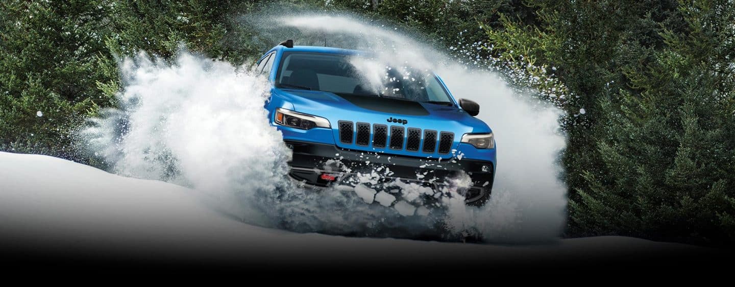 A blue 2023 Jeep Cherokee Trailhawk bursting through a mound of snow with white powder billowing over the sides and the top of the vehicle and evergreens in the background.
