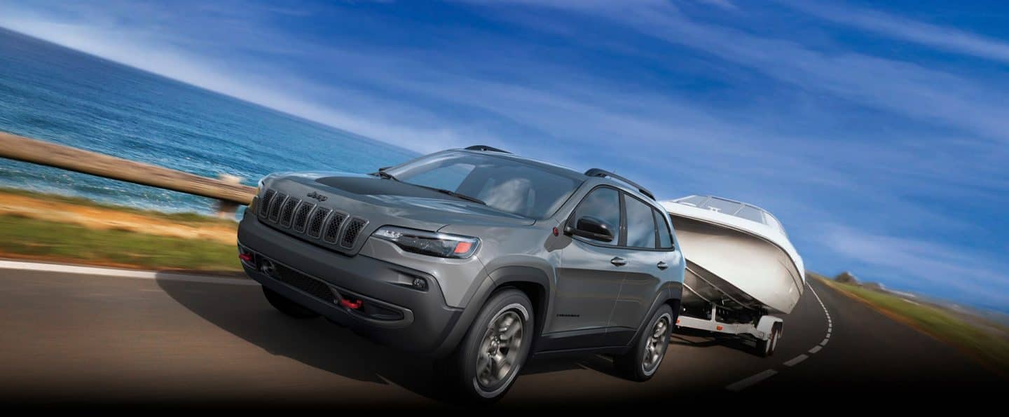 A gray 2023 Jeep Cherokee Trailhawk being driven along a seaside highway, towing a motorboat.