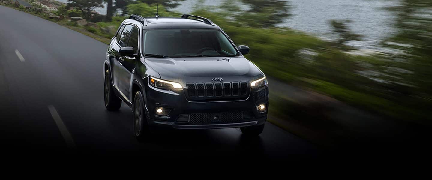 A 2023 Jeep Cherokee Altitude Lux being driven on road beside a lake with an overcast sky and the background blurred to indicate the vehicle is in motion.