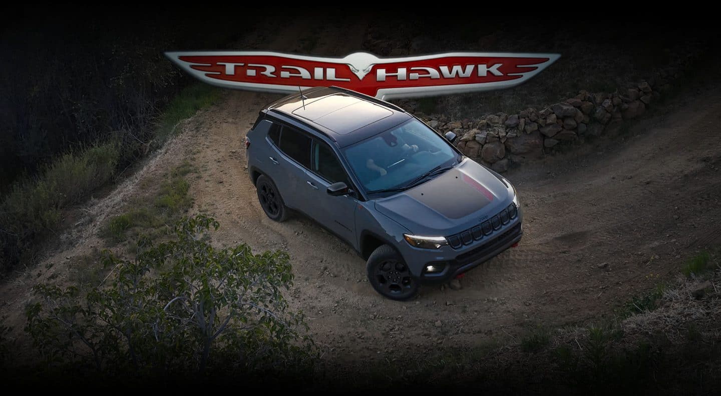 Un Jeep Compass Trailhawk 2023 andando por una colina sin asfaltar junto a una cerca de piedra. El emblema de Trailhawk.