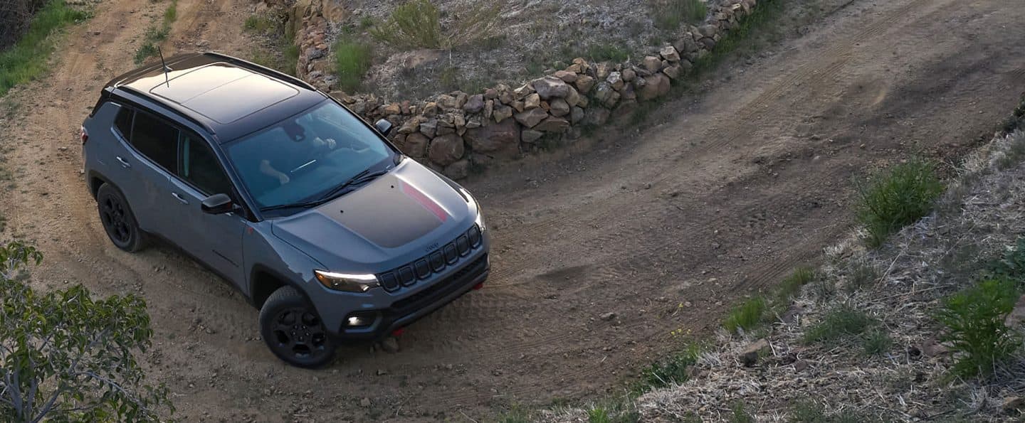 A 2023 Jeep Compass Trailhawk being driven on a dirt road hill beside a stone fence.