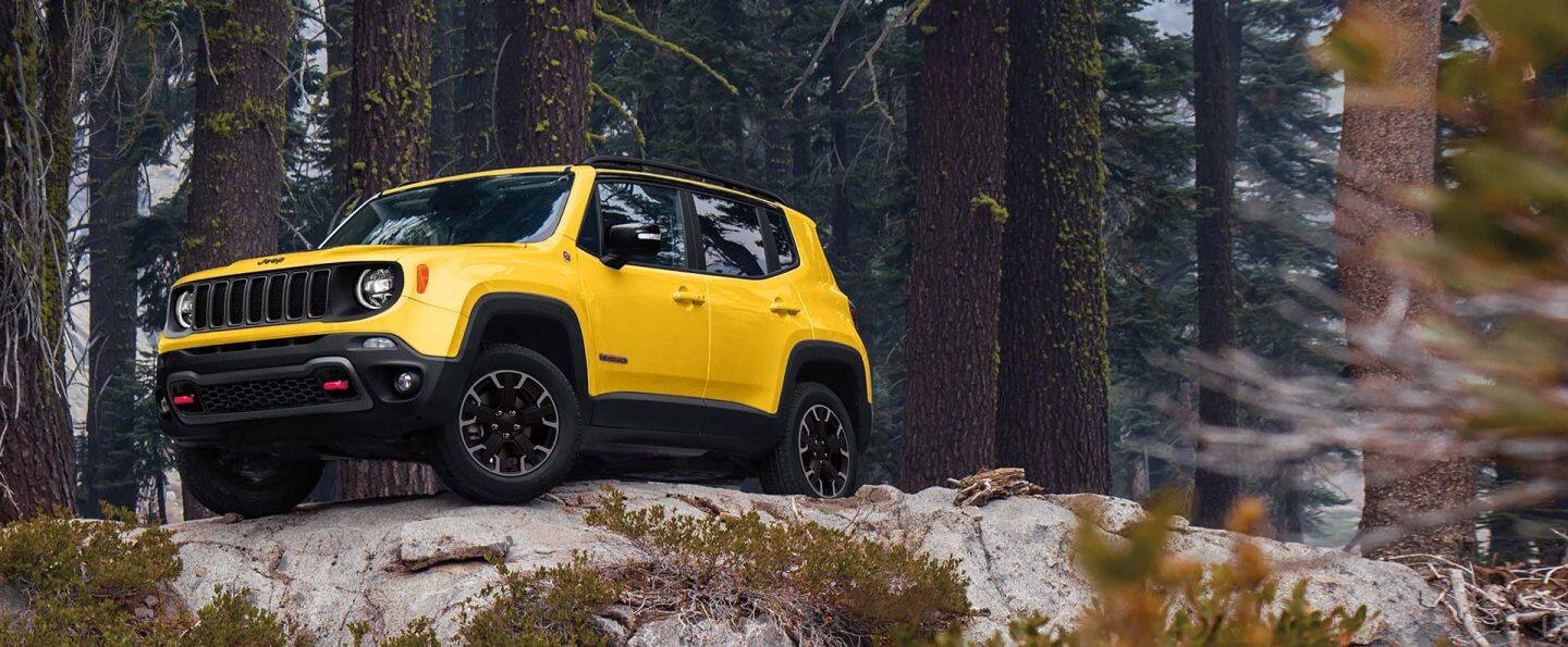 A yellow 2023 Jeep Renegade Trailhawk parked on a rocky outcrop in a wooded area off-road.