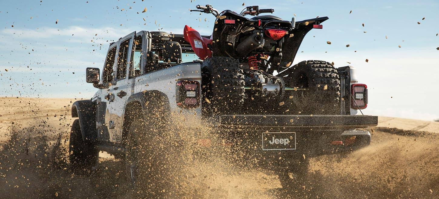2023 Jeep Gladiator Rubicon toz buludunda yolsuzluqda sürülür, yük maşınının çarpayısına dörd təkərli avtomobili yerləşdirmək üçün yük qapısı aşağı salınır.