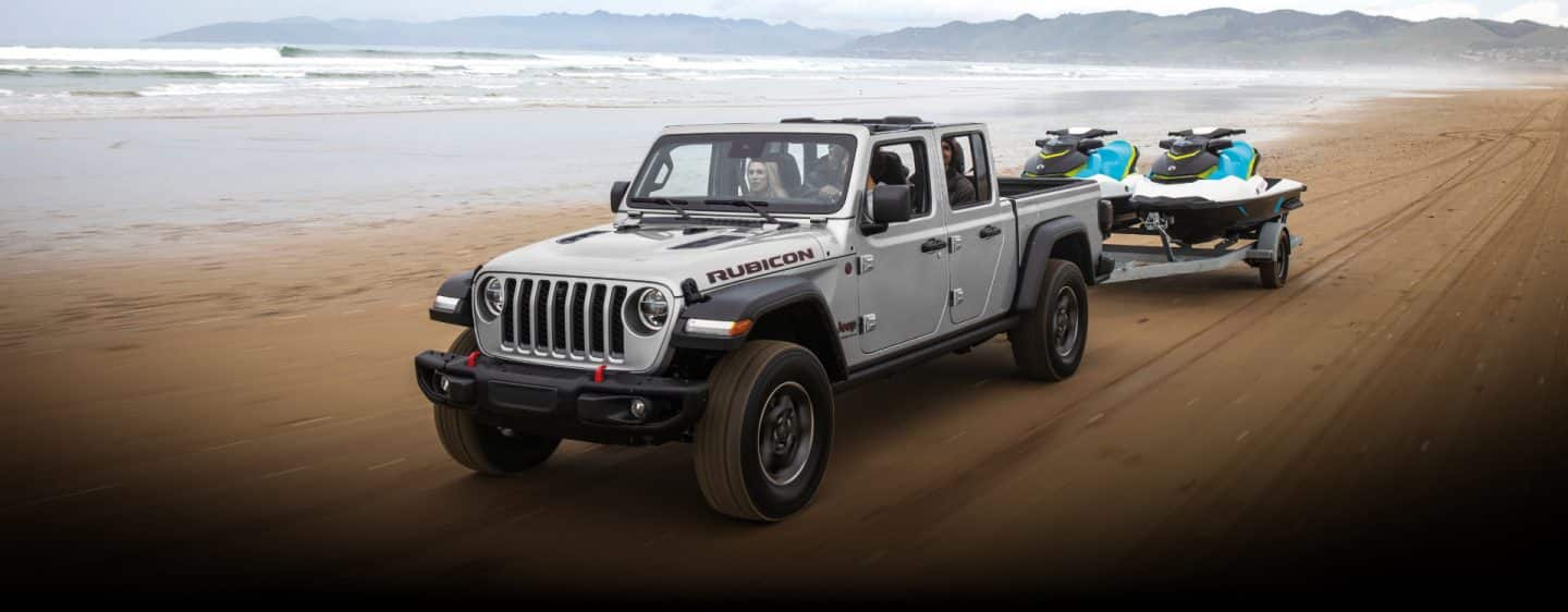 A 2023 Jeep Gladiator Rubicon towing two jet skis as it is driven along a beach near the water.