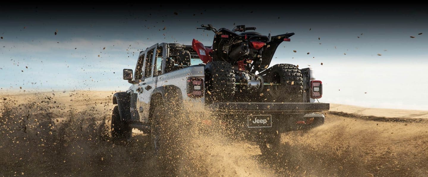 Vista trasera de una Jeep Gladiator Rubicon 2023 levantando arena a medida que avanza por un desierto, llevando un ATV en su plataforma con la puerta trasera abierta.