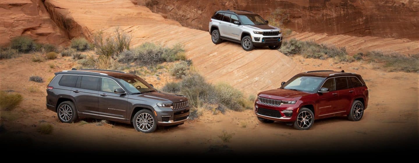 Three 2023 Jeep Grand Cherokee models, including the three-row Grand Cherokee L Summit Reserve and the Grand Cherokee 4xe, parked off-road in scrubby desert terrain.