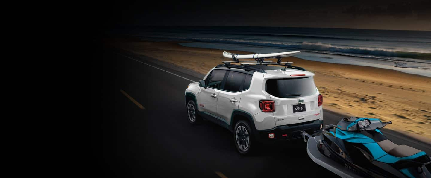 Un Jeep Renegade Trailhawk 2023 blanco con una tabla de surf en la parrilla portaequipaje, remolcando una moto de agua.