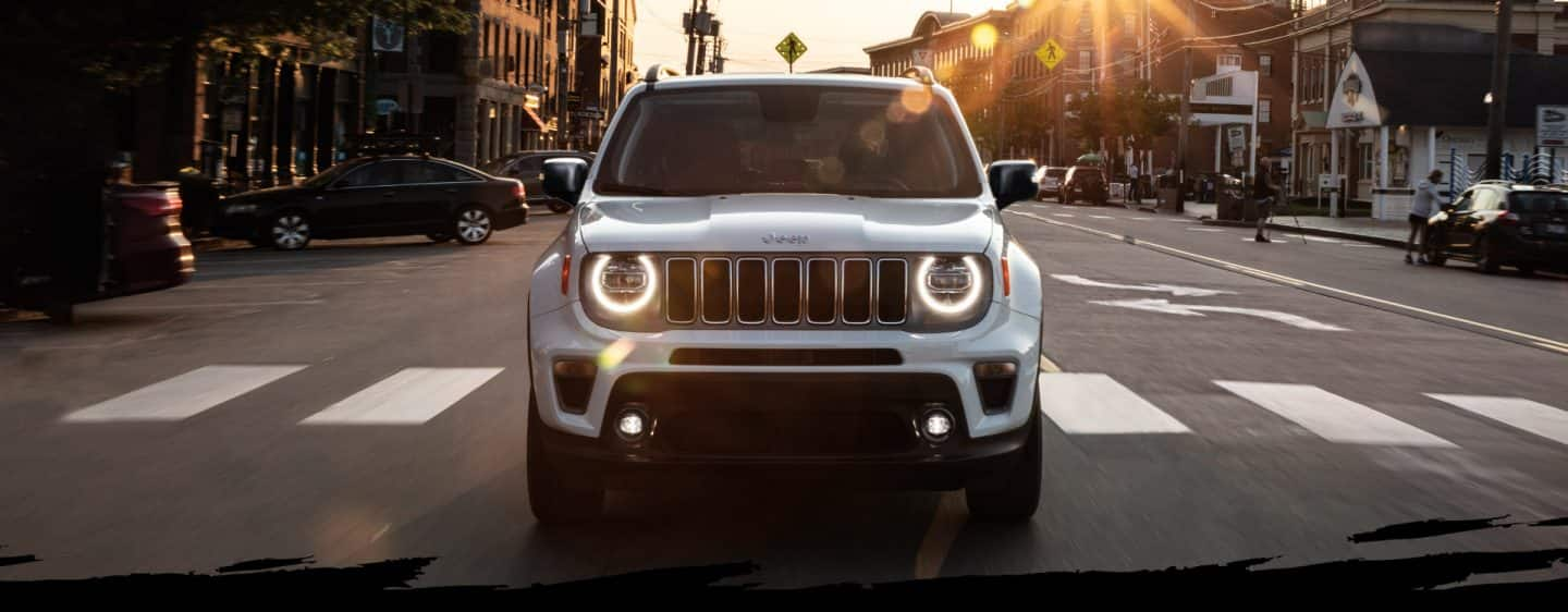 Una vista frontal del Jeep Renegade Limited 2023 circulando con los faros delanteros encendidos por una concurrida calle de la ciudad al atardecer.