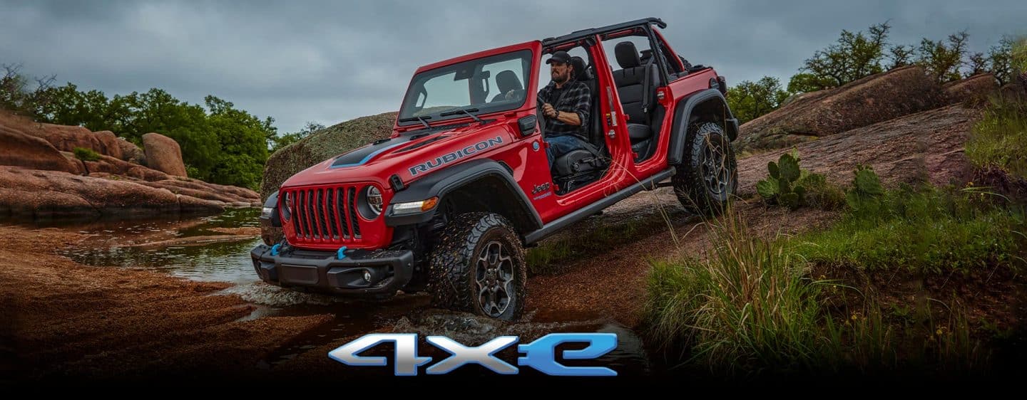 A gray 2023 Jeep Wrangler Willys 4xe parked on a rocky outcropping with a stormy sky above and a forest of trees below. The Memorial Day Sales Event logo.