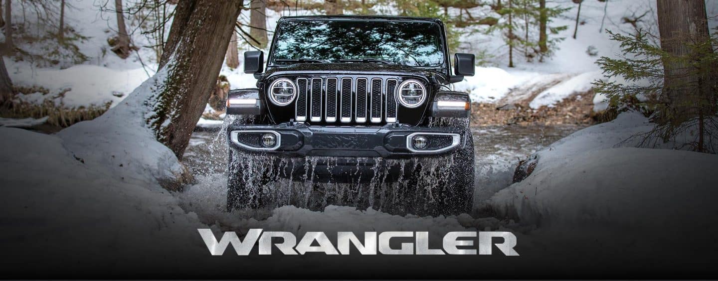 A head-on view of a 2023 Jeep Wrangler Sahara climbing out of an icy stream onto the snow, in a forest off-road. Wrangler.