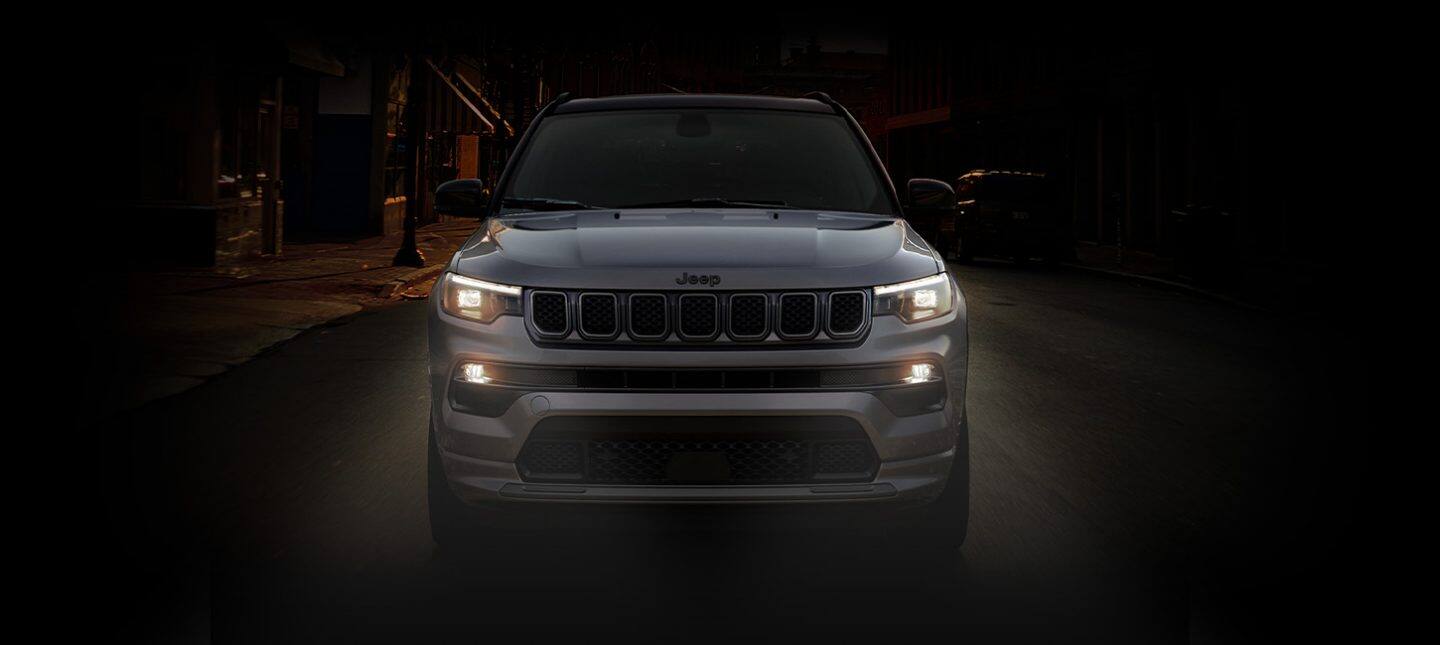 Un unghi frontal al unui Jeep Compass 2024 High Altitude condus pe o stradă din oraș pe timp de noapte, cu farurile și lămpile de ceață aprinse.