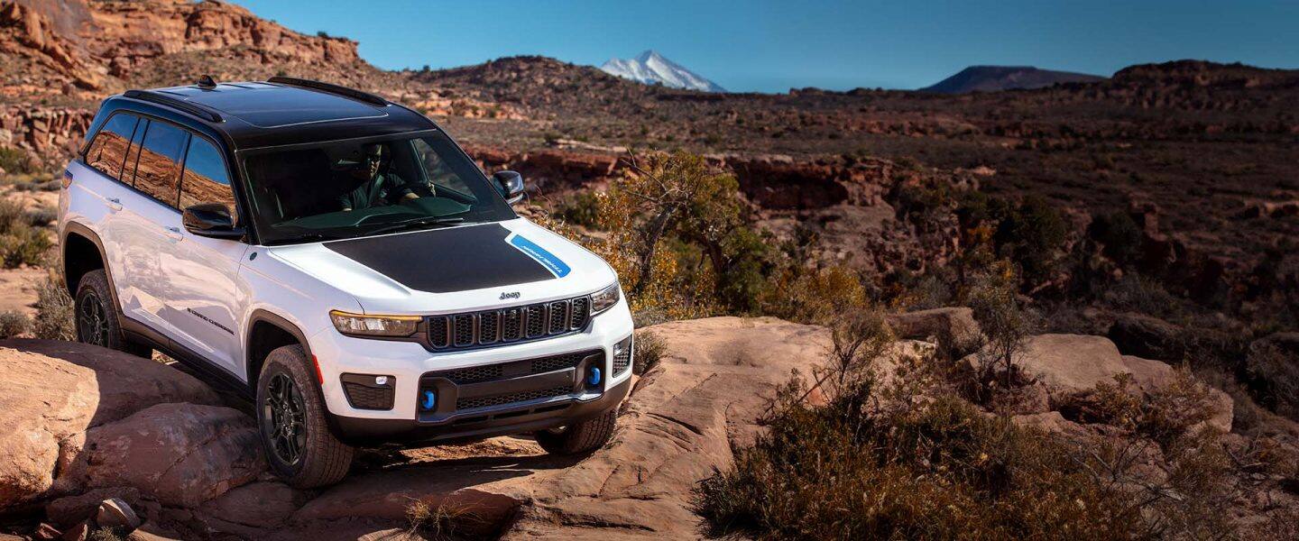 Un Jeep Grand Cherokee 4xe Trailhawk alb din 2024 care se târa peste bolovani mari în deșert, cu munți în fundal.