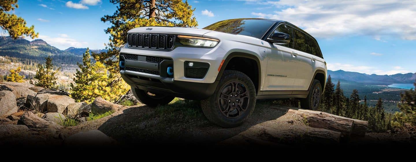 Un Jeep Grand Cherokee Trailhawk 4xe 2024 blanco con una inserción negra en el capó, circulando por un afloramiento rocoso, con montañas y un lago de fondo.