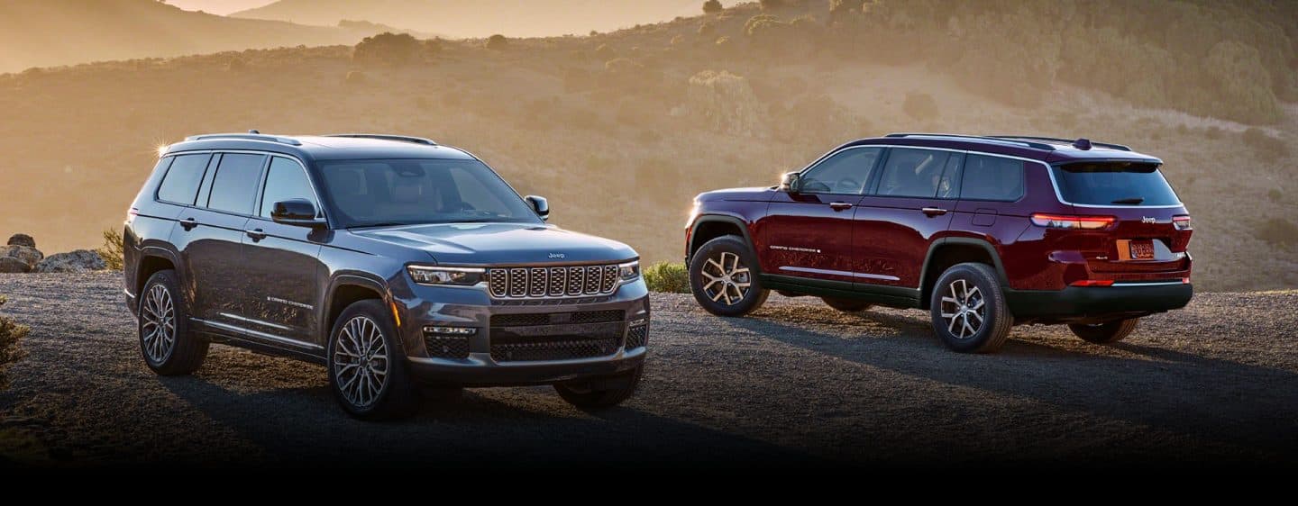 Un Jeep Grand Cherokee Summit Reserve gris y un Jeep Grand Cherokee Limited 2024 rojo estacionado uno frente al otro en una carretera de montaña de ripio al atardecer.