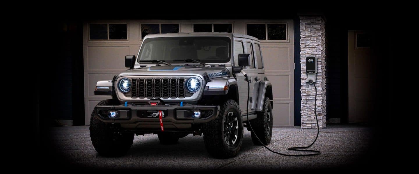 Un Jeep Wrangler Rubicon 4xe estacionado en un garaje con una estación de carga en la pared y el cable de carga enchufado al puerto de carga del vehículo.