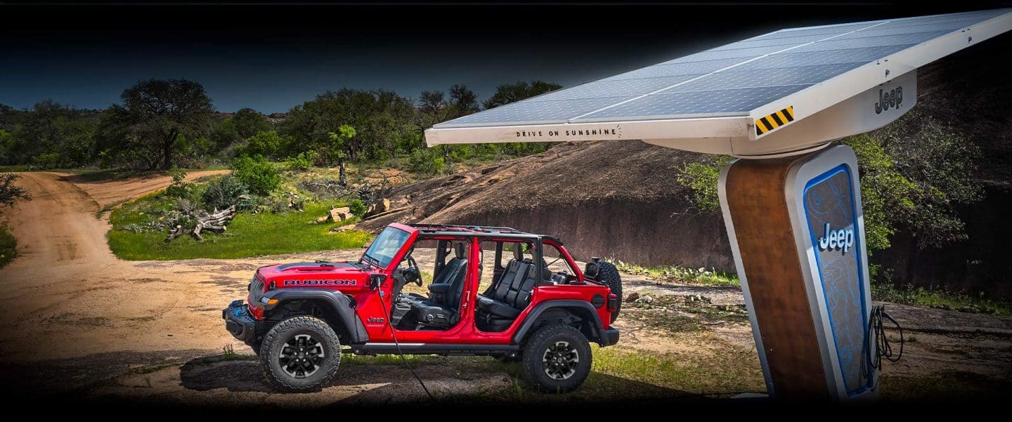 Un Jeep Wrangler Rubicon 4xe 2024 rojo, con las puertas y el techo desmontados, se muestra estacionado junto a una estación de carga con alimentación solar de la marca Jeep. El cable de carga está enchufado al puerto de carga del vehículo.