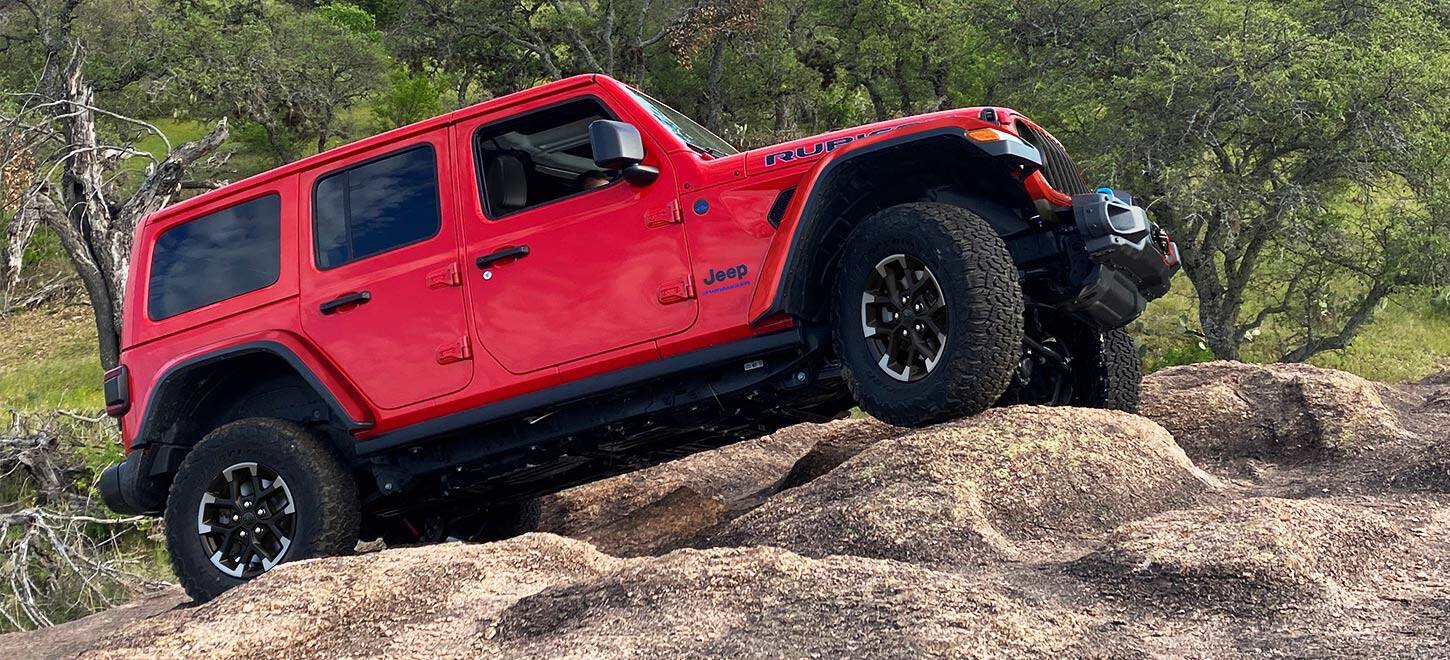 Профиль красного Rubicon 2024 Jeep Wrangler 4xe со стороны пассажира, поднимающийся на холм покрытых песком валунов.