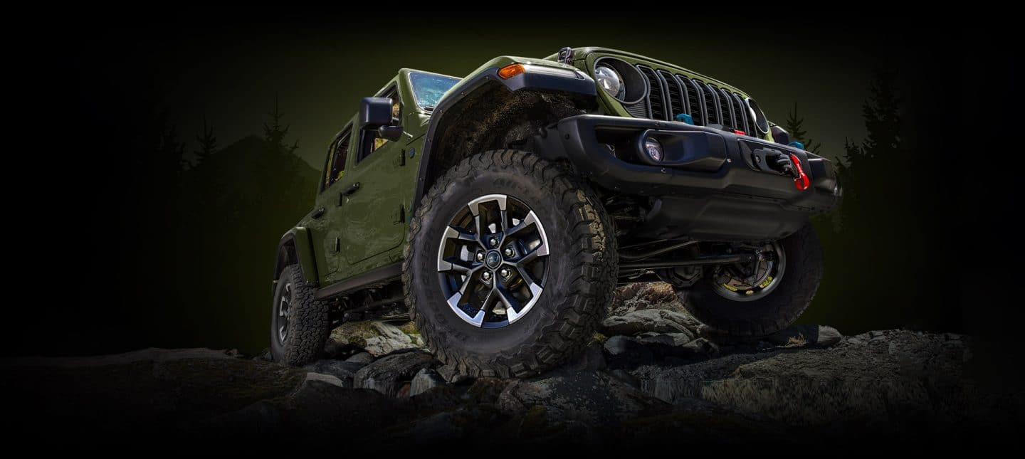 A angle from below of a green 2024 Jeep Wrangler Rubicon 4xe crawling over a stoney mound off-road.