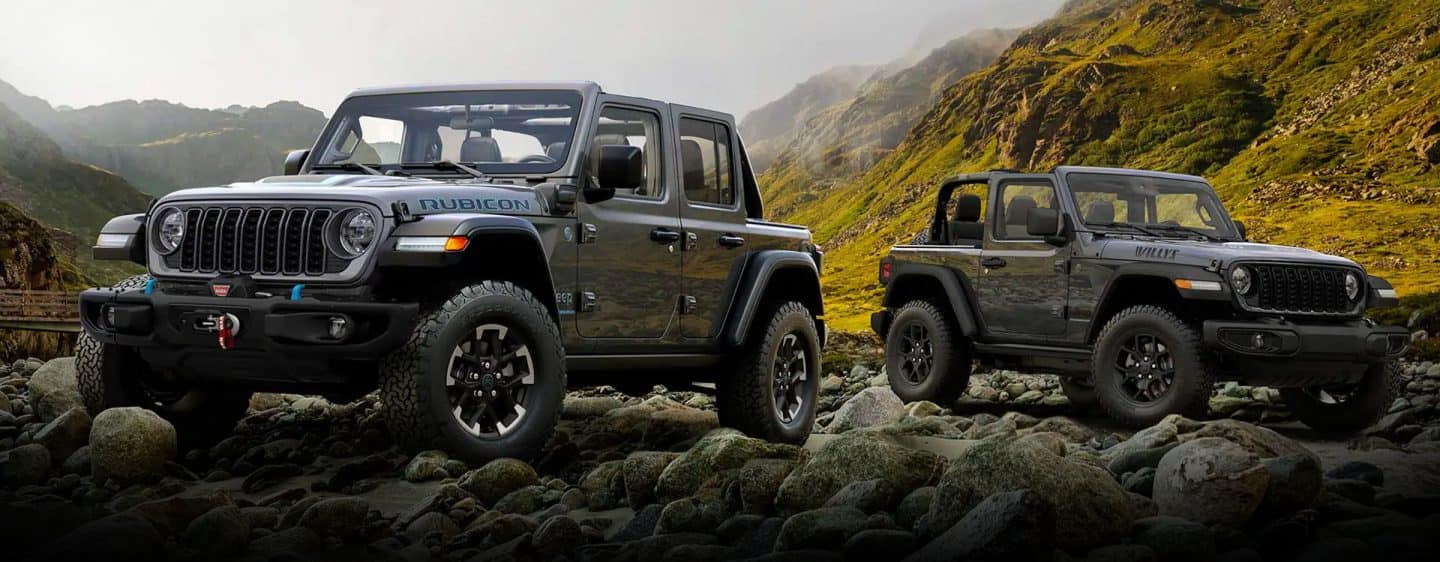 Dos modelos de Jeep Wrangler estacionados en un claro rocoso en las montañas. A la izquierda, un Wrangler 4xe Rubicon gris de cuatro puertas. A la derecha, un Wrangler Willys gris de dos puertas.