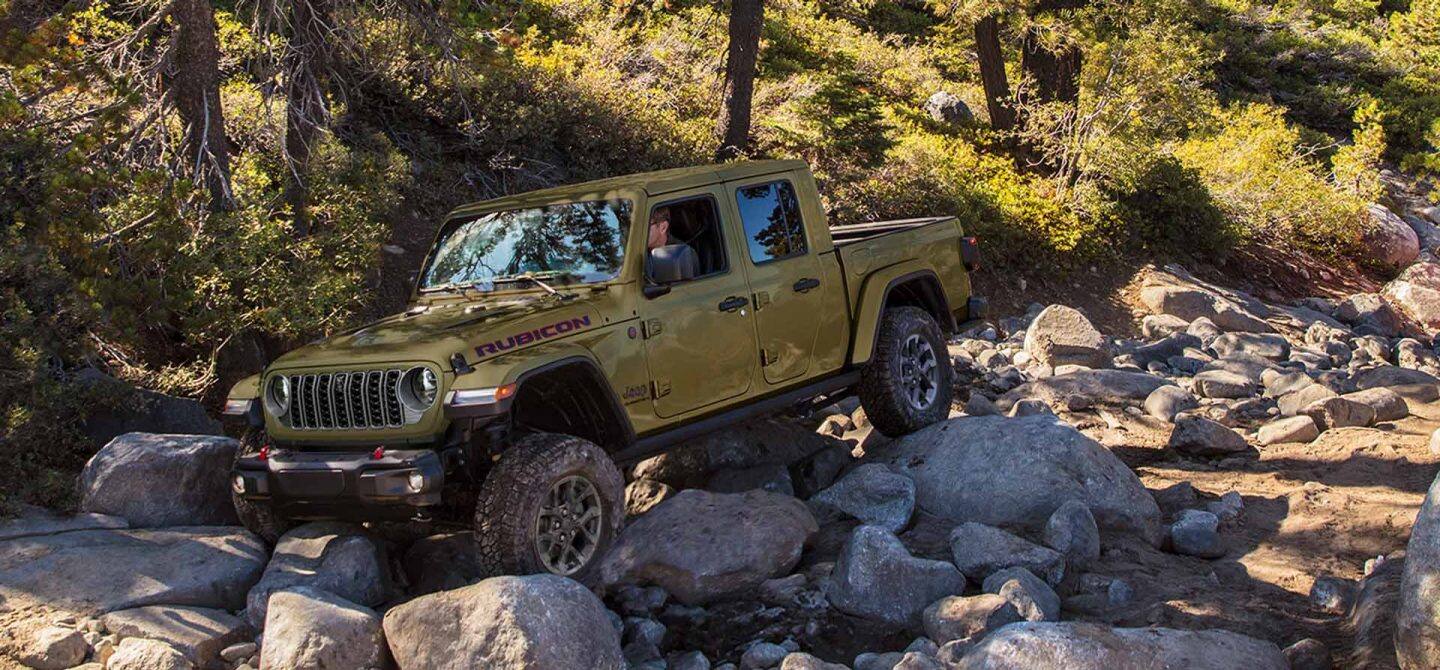 Un Jeep Gladiator Rubicon X verde oliva de 2025 sobre una serie de rocas en un bosque todoterreno.