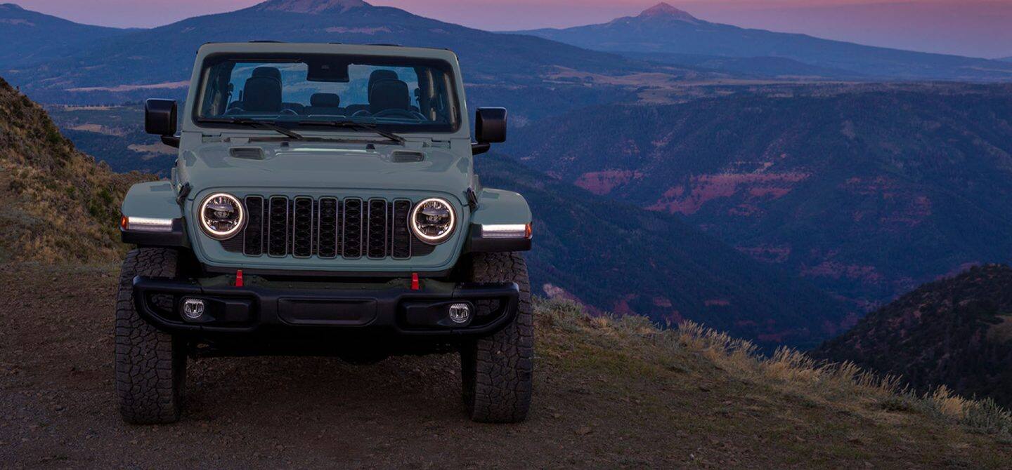 La parte delantera de un Jeep Gladiator Rubicon X gris de 2025 aparcado en el borde de un acantilado con montañas a la distancia.