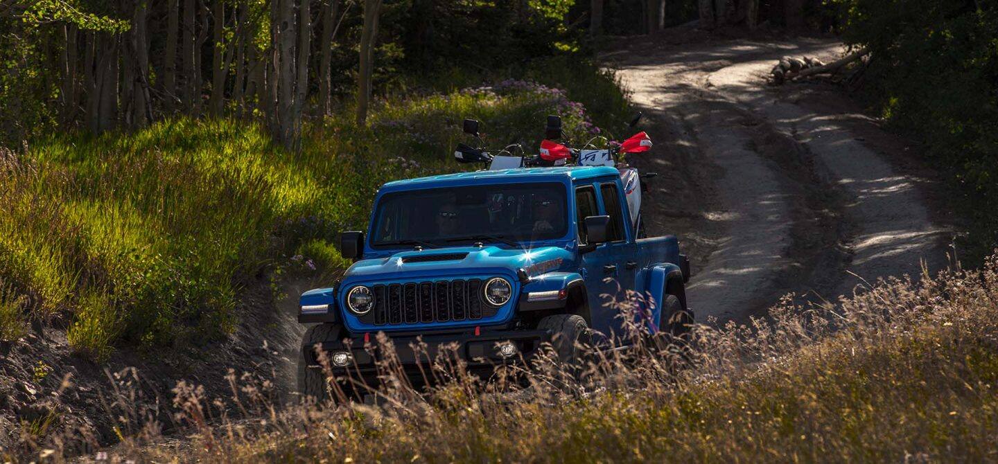 Un Jeep Gladiator Mojave X 2025 azul que se conduce por un sendero sinuoso con dos ATV en su caja de recogida.