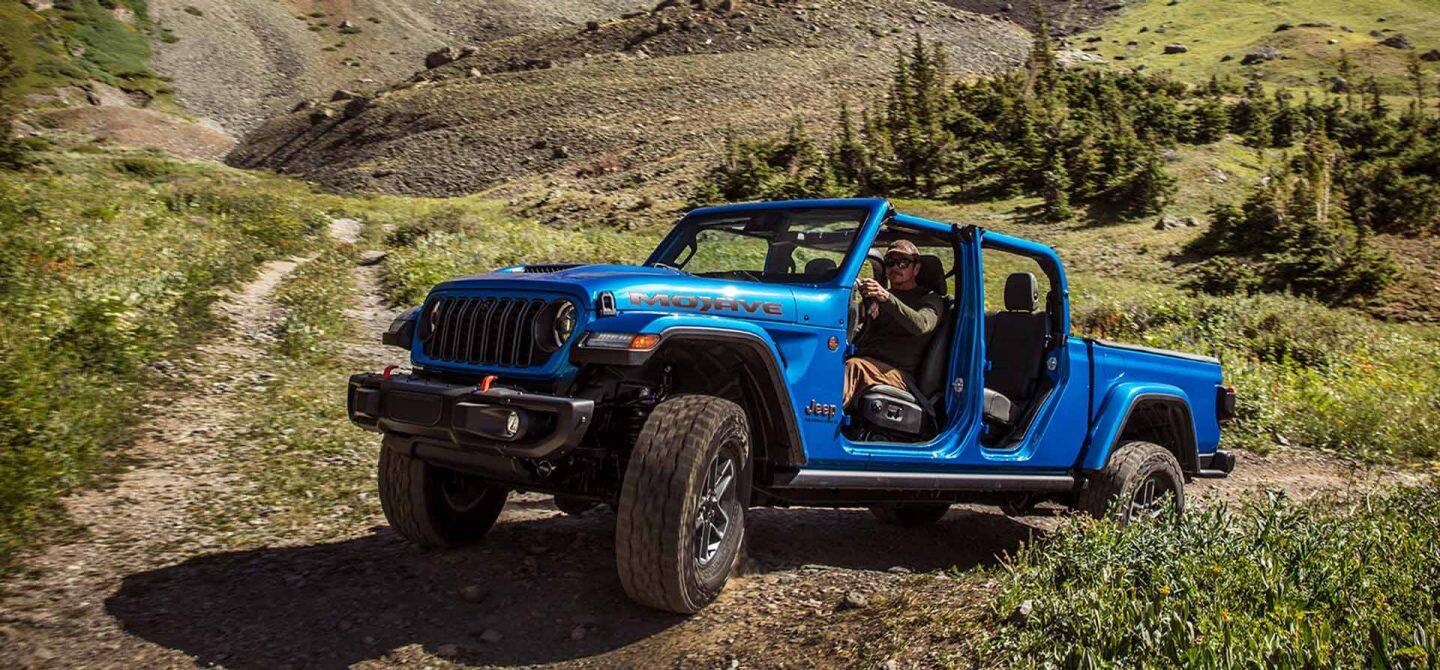 Un Jeep Gladiator Mojave X 2025 azul con sus puertas y techo retirados, ascendiendo por un sendero en una zona montañosa todoterreno.