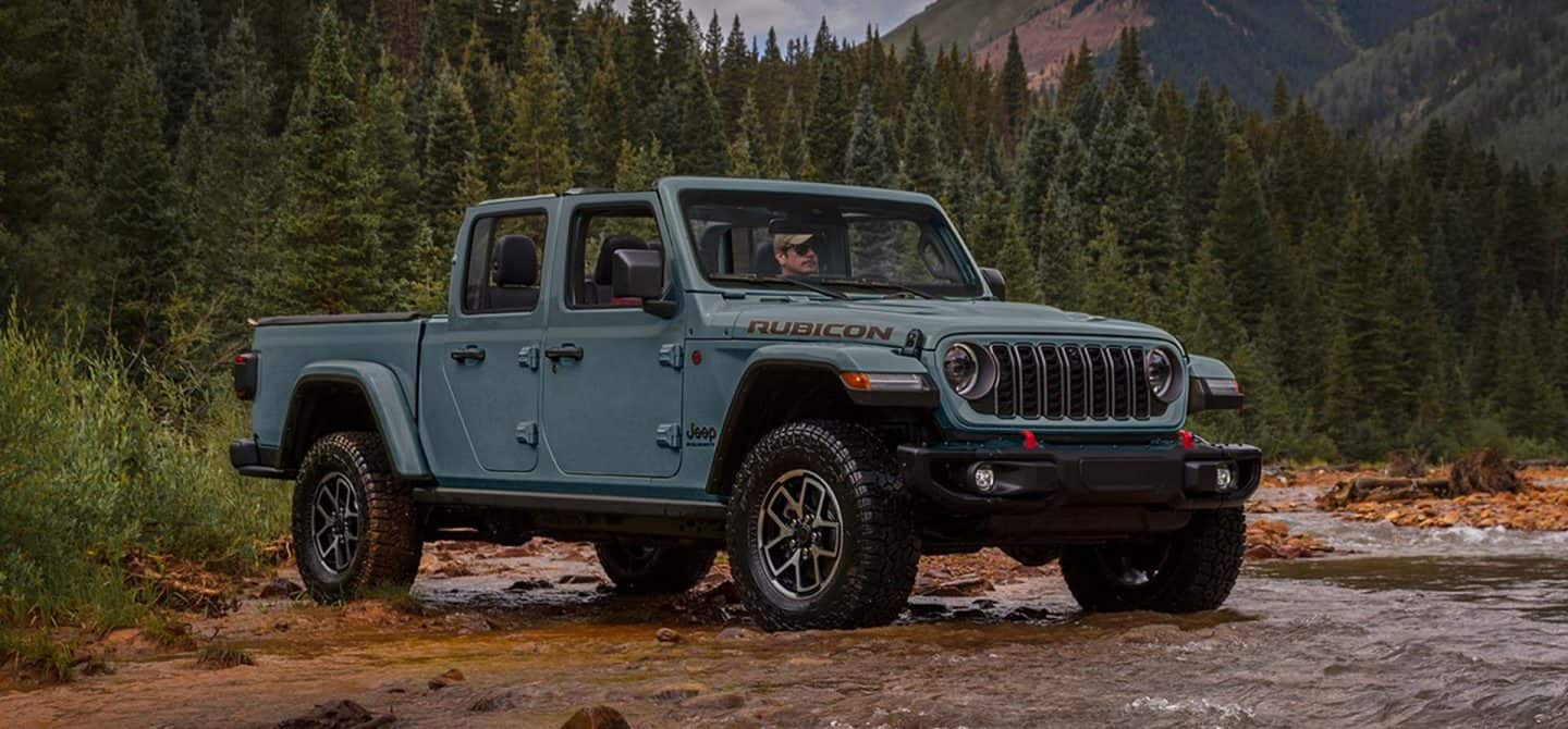 Un Jeep Gladiator Mojave X 2025 azul con sus puertas y techo retirados, ascendiendo por un sendero en una zona montañosa todoterreno.