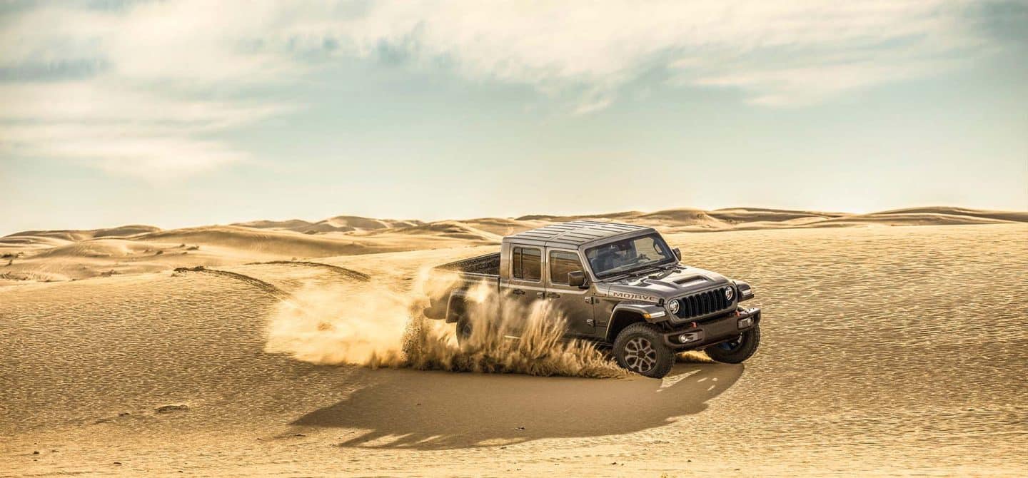 Un Jeep Gladiator Mojave X gris de 2025 que levanta la arena mientras se conduce por una llanura del desierto.