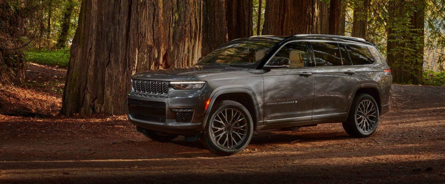 Ángulo frontal de un Jeep Grand Cherokee Trailhawk 4xe 2025 plateado con una inserción negra en el capó, circulando por un camino de tierra en las montañas, cerca de un cuerpo de agua.