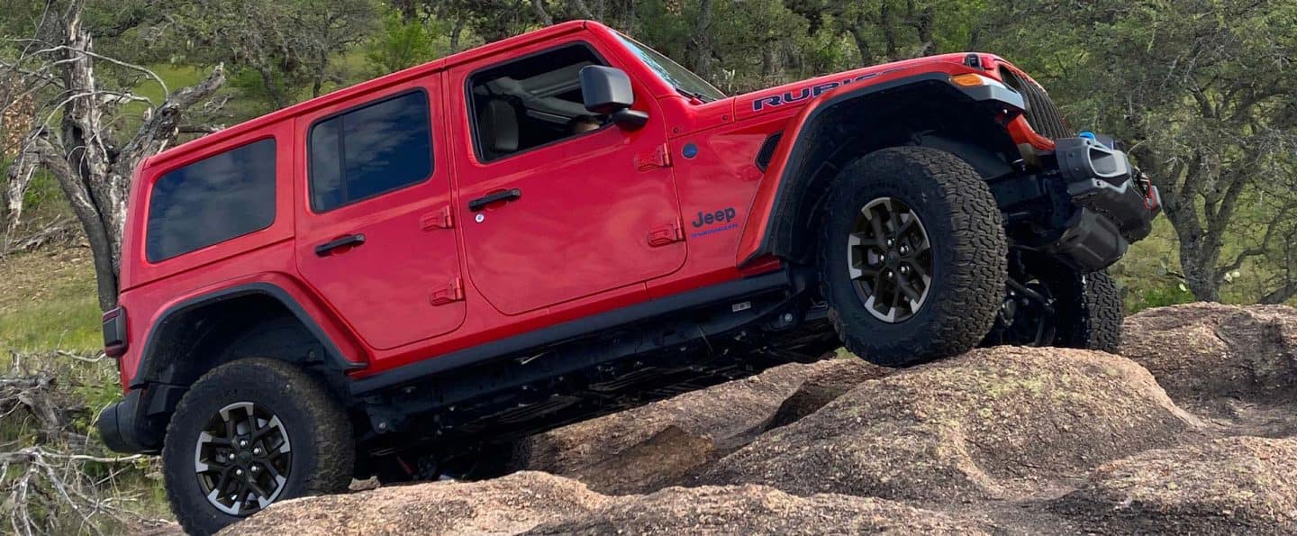 Un perfil del lado del pasajero de un Jeep Wrangler Rubicon X 4xe rojo 2025 escalando una colina de rocas cubiertas de arena.