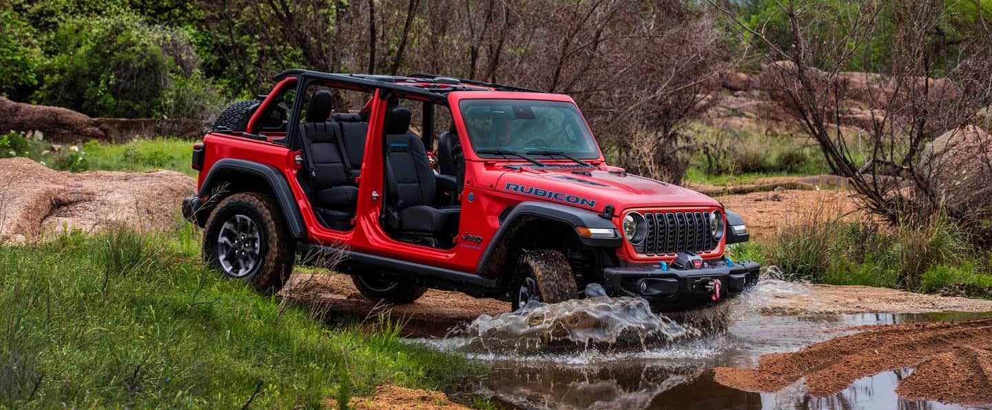Un Jeep Wrangler Rubicon 4xe rojo 2025 con sus puertas y techo retirados, entrando en un arroyo todoterreno.
