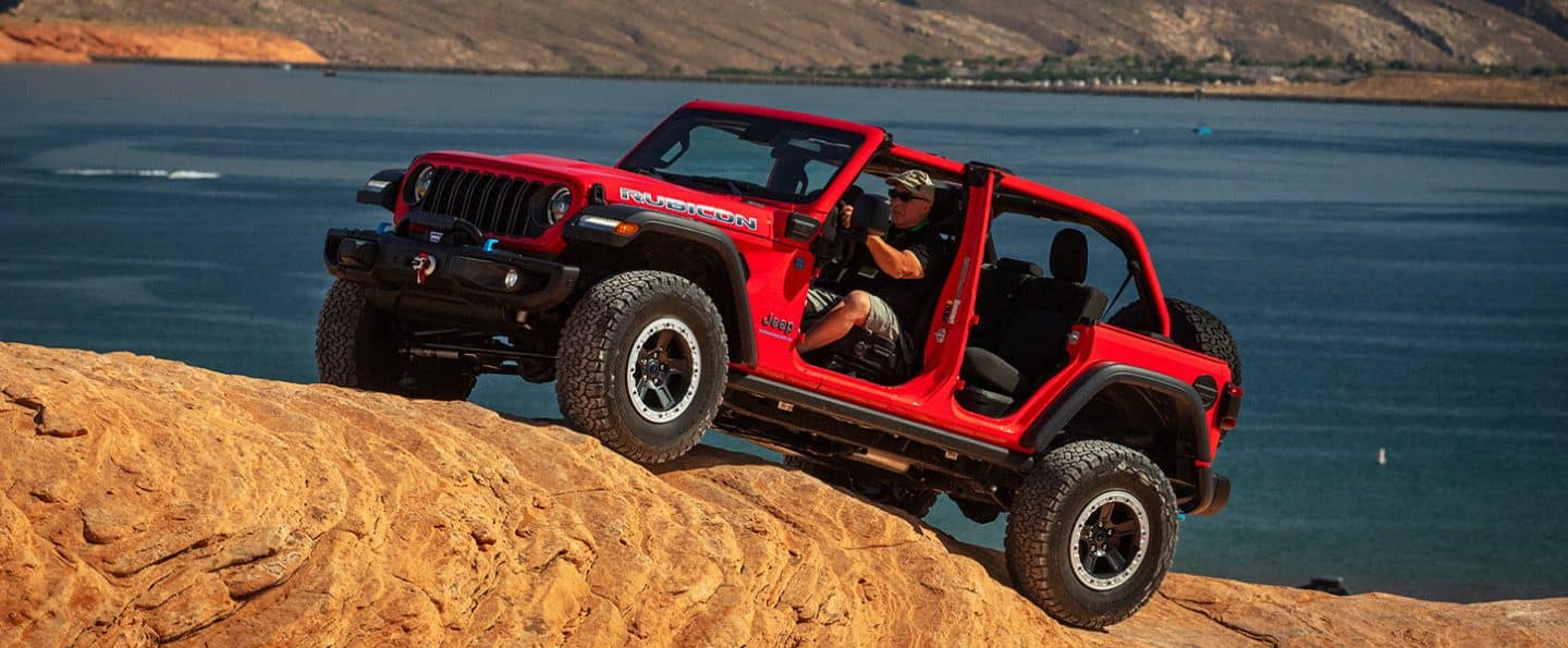 Un Jeep Wrangler Rubicon 4xe rojo de 2025 con sus puertas y techo retirados, ascendiendo por una colina arenosa en una playa.