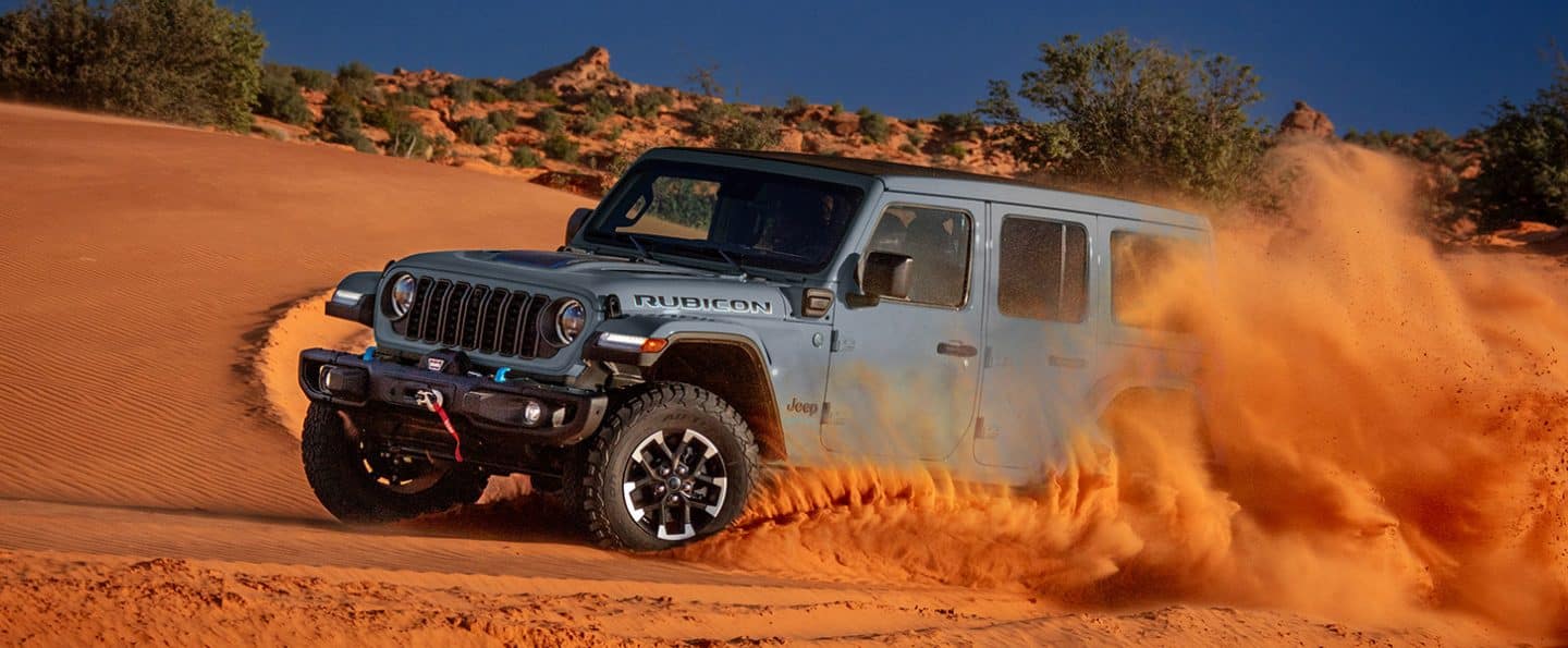 Un Jeep Wrangler Rubicon X 4xe gris de 2025 que viaja a través de una duna en el desierto, arrasando arena sobre la marcha.
