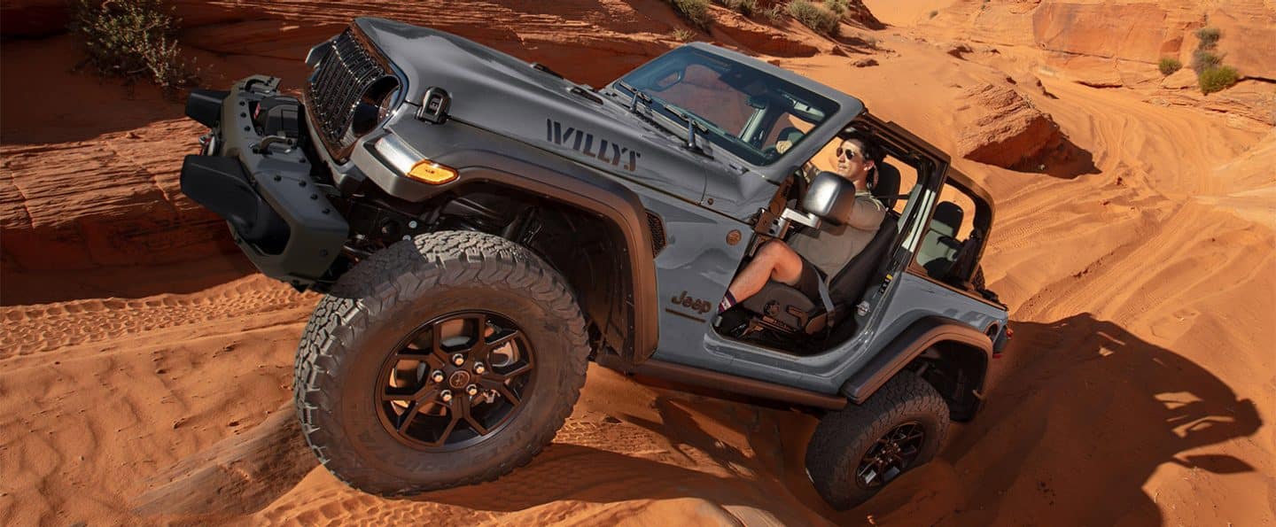 Un Jeep Wrangler Willys gris de 2025 con sus puertas y techo retirados, gateando y sobre una serie de dunas de arena en el desierto.