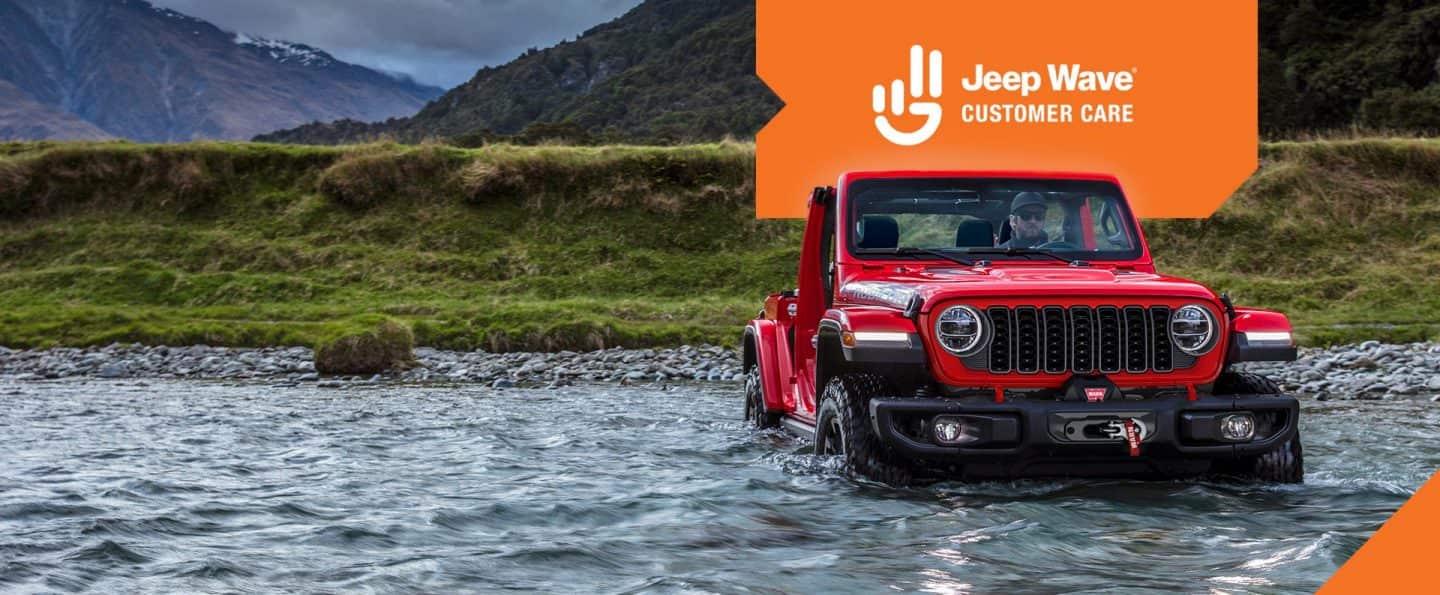 Un ángulo frontal de un Jeep Wrangler Rubicon X rojo 2025 forzando un arroyo con el agua a mitad de camino hacia las ruedas. Jeep Logotipo de Wave Customer Care.