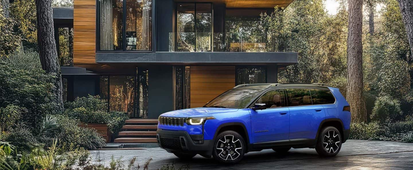 Un Jeep Cherokee Overland 2026 azul estacionado en la entrada de una gran casa contemporánea en el bosque.