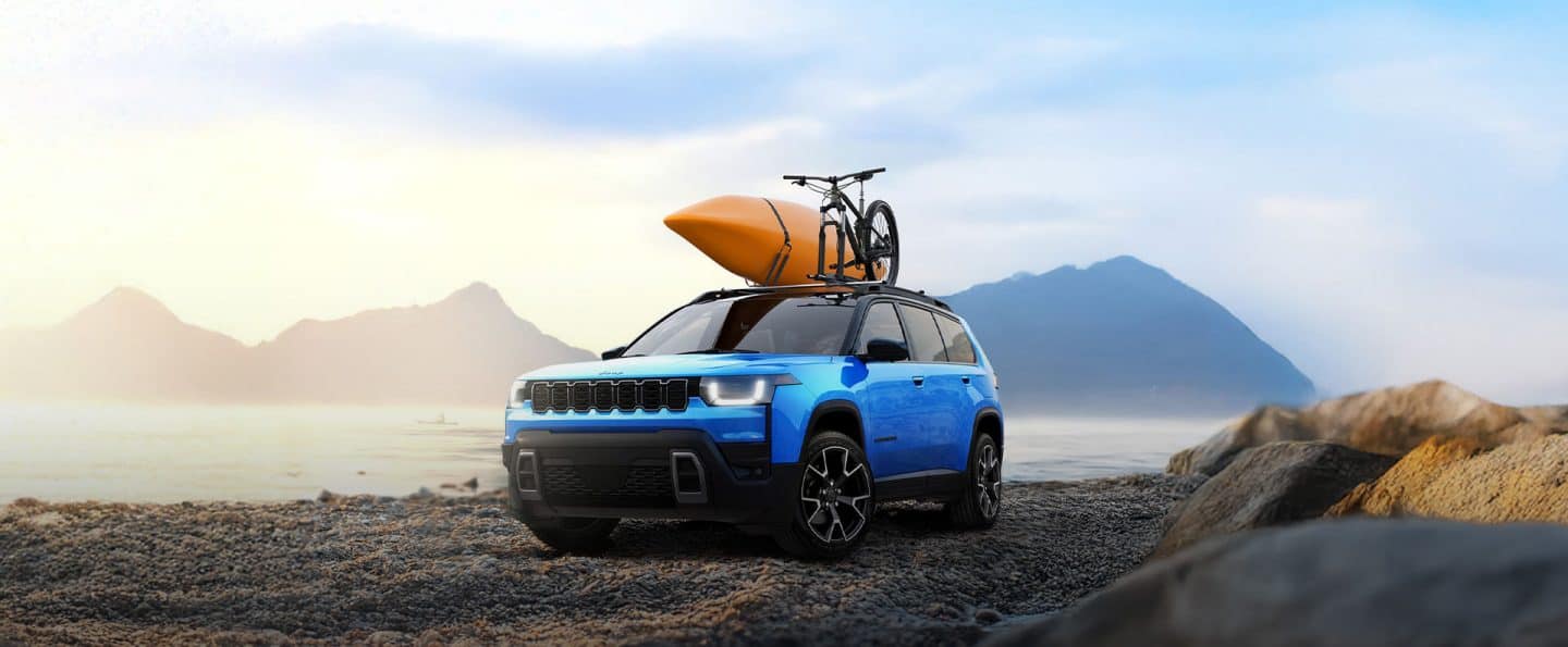 Un Jeep Cherokee Overland 2026 azul estacionado en una playa rocosa con un kayak y parte de una bicicleta amarrada a la parrilla portaequipaje.