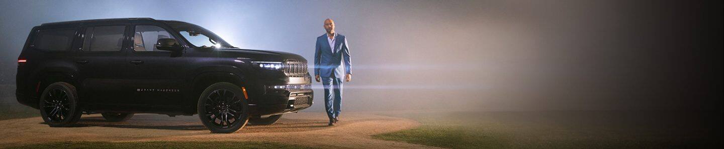 Un perfil de la Grand Wagoneer Obsidian 2022 con los faros delanteros encendidos, estacionada sobre el montículo del lanzador de un campo de béisbol por la noche, con una luz que se abre camino entre un fondo con niebla. Derek Jeter, caminando delante del vehículo.