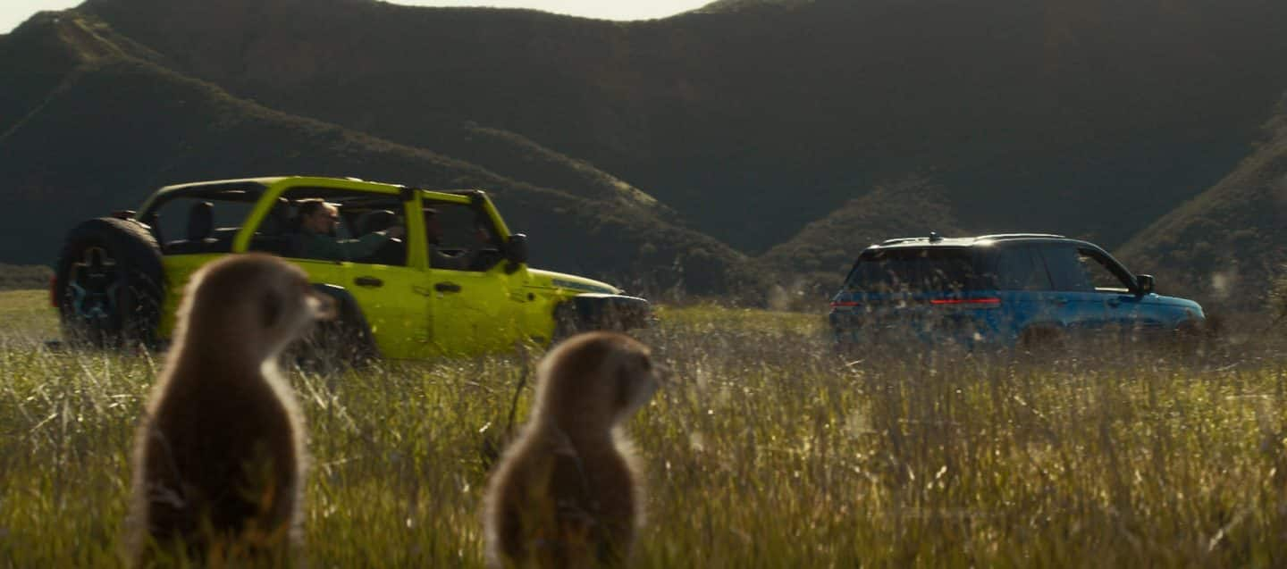 A white 2023 Jeep Wrangler Rubicon 4xe and a lime green 2023 Jeep Gladiator Rubicon, both with their doors and roofs removed, parked opposite of each other on sand at the foot of a large, rocky hill. Memorial Day Sales Event.