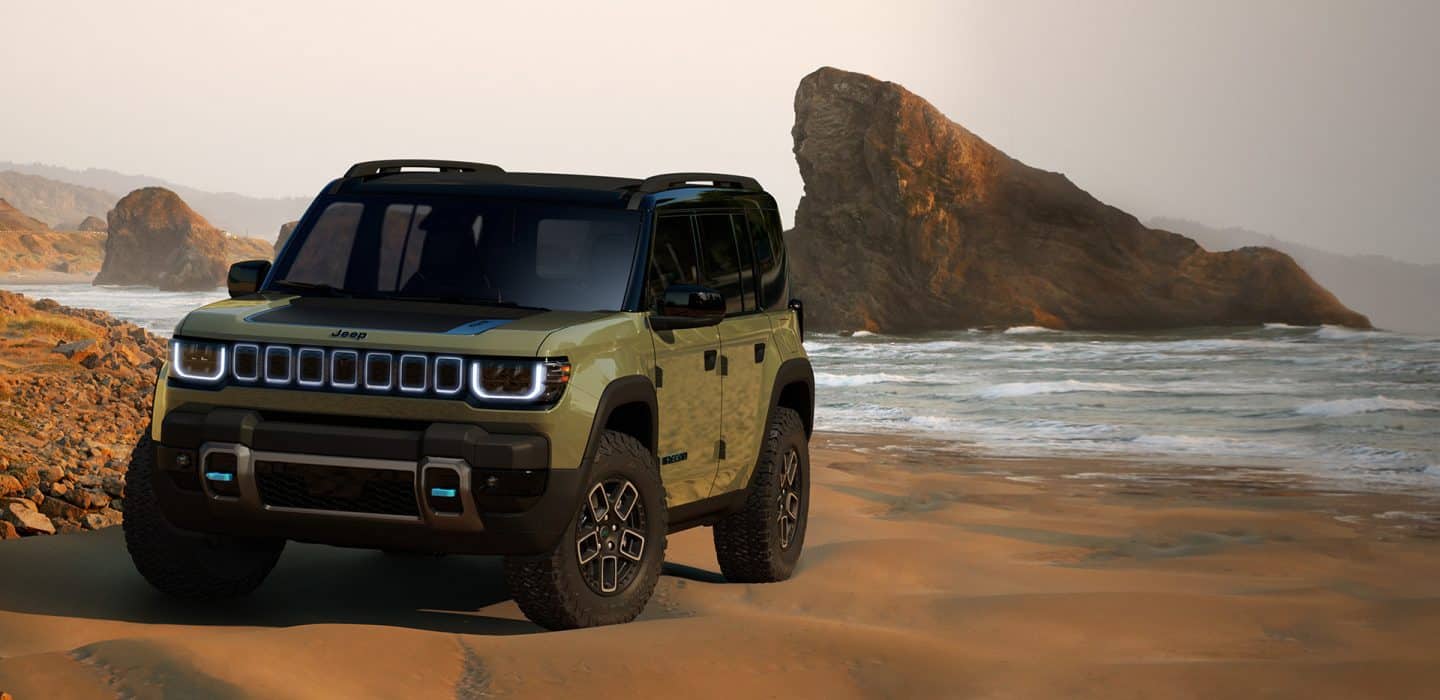 El modelo en preproducción de Jeep Recon 4xe estacionado en una playa de arena roja con grandes formaciones rocosas a la distancia.