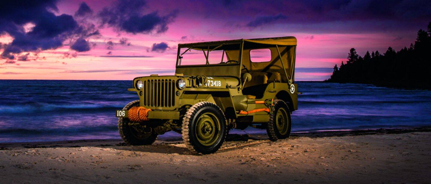 Une Seconde Guerre mondiale Jeep garée sur une plage au coucher du soleil, avec un ciel rose et violet en arrière-plan.