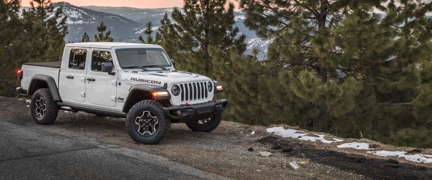 A profile of a white 2023 Jeep Gladiator Rubicon parked on the edge of a dropoff, with evergreens and mountains in the background.