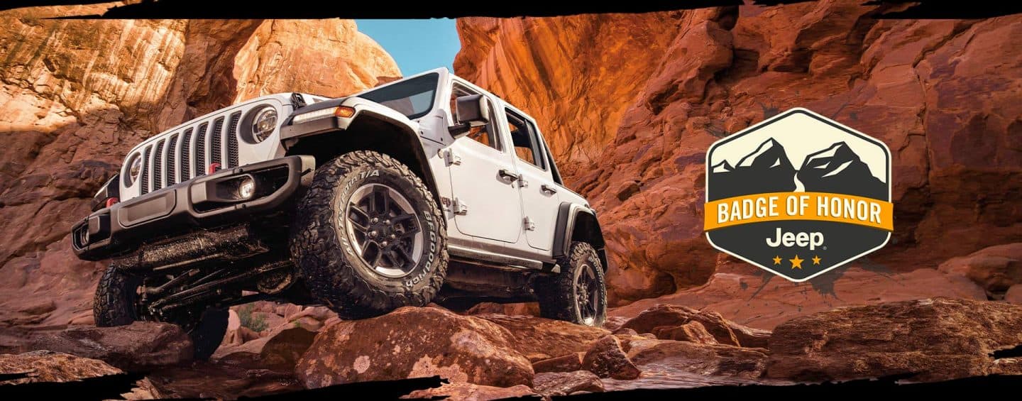 A 2023 Jeep Wrangler Rubicon, seen from below as it surmounts a rock in a canyon. Jeep Badge of Honor. 