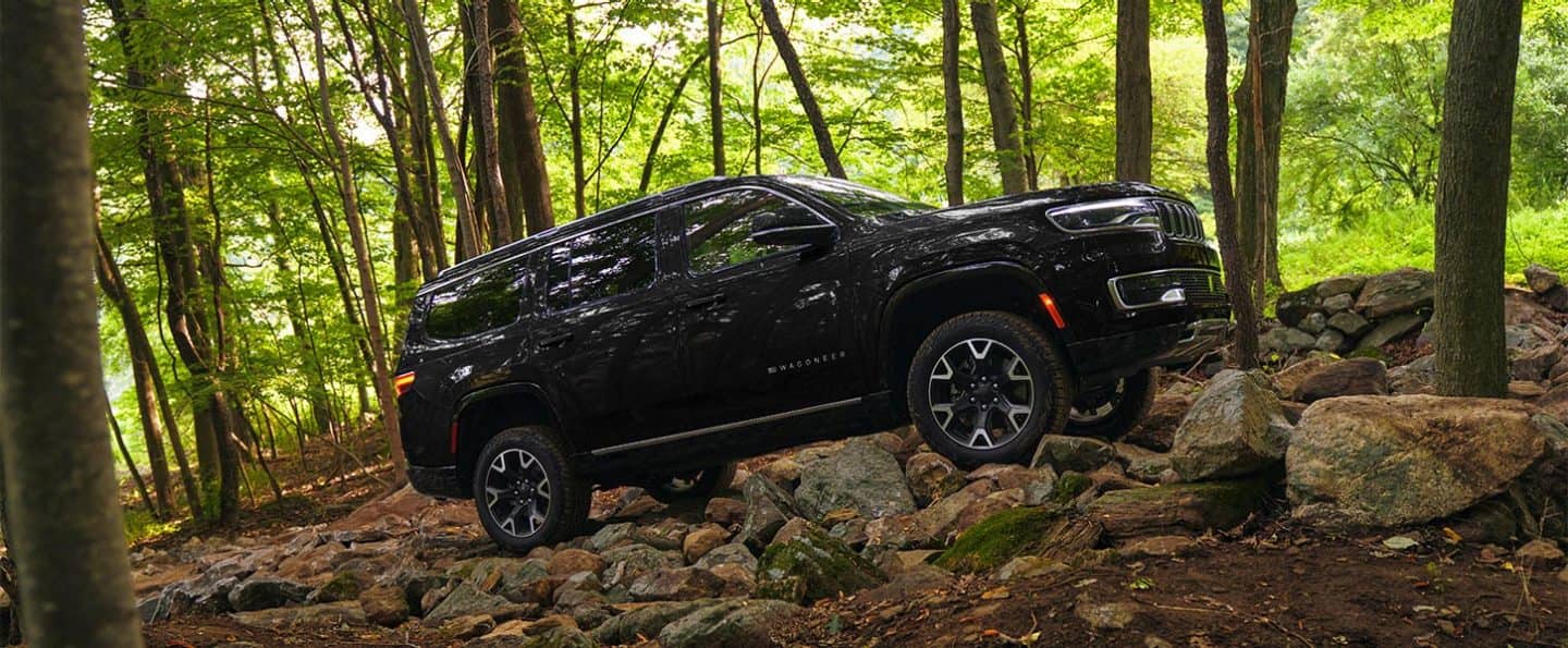 Perfil del lado del pasajero de un Jeep Wagoneer Series 3 2025 negro trepando una colina rocosa por el bosque. 