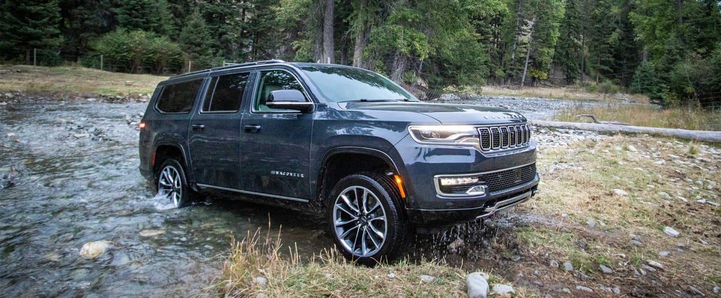 Un ángulo del lado del pasajero de un Jeep Wagoneer Serie 3 azul del año 2025 saliendo de un arroyo en un sendero todoterreno en el bosque.