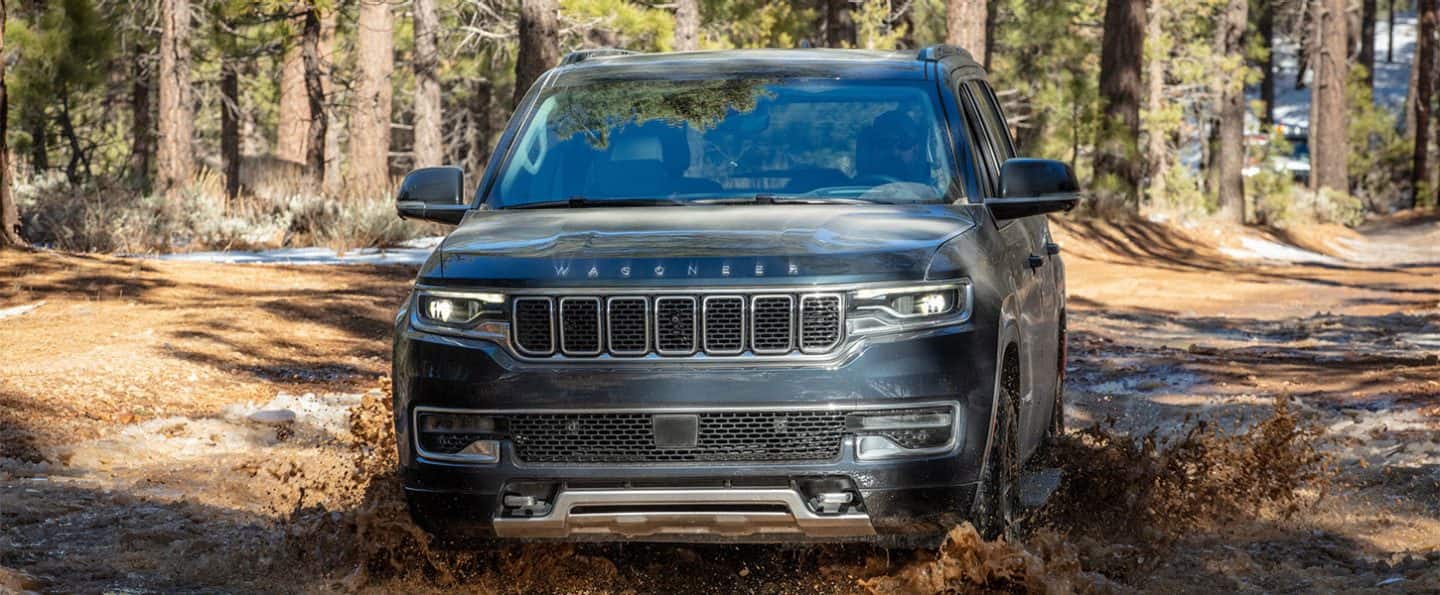 Ángulo superior de un Jeep Wagoneer Series 3 2025 gris vadeando un arroyo poco profundo lleno de lodo en un sendero en el bosque.    